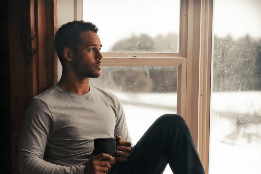 Man sitting by a snowy window holding a warm mug, pausing to reflect on what it means to be human
