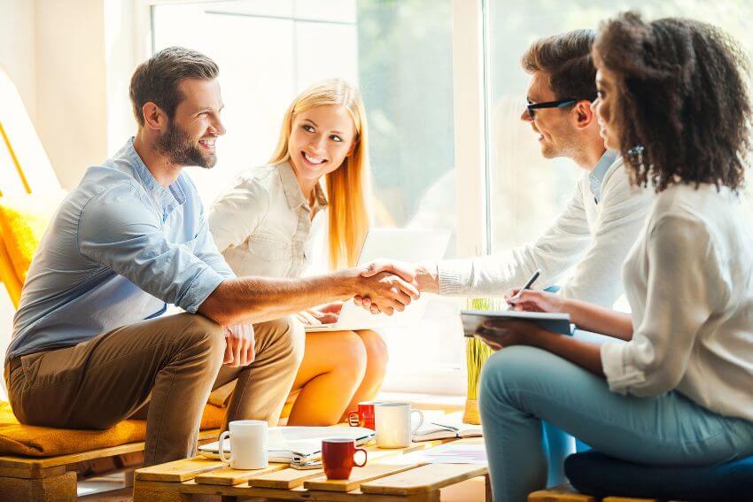 Four young colleagues sharing a warm handshake in a sunlit room, a reminder of how much it matters to surround yourself with good people who show up genuinely.