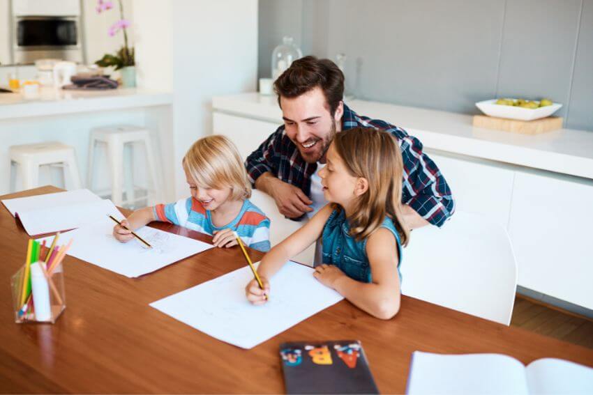 A father leaning in and laughing with his two children as they draw at the kitchen table, showing up fully with time and attention that costs nothing.