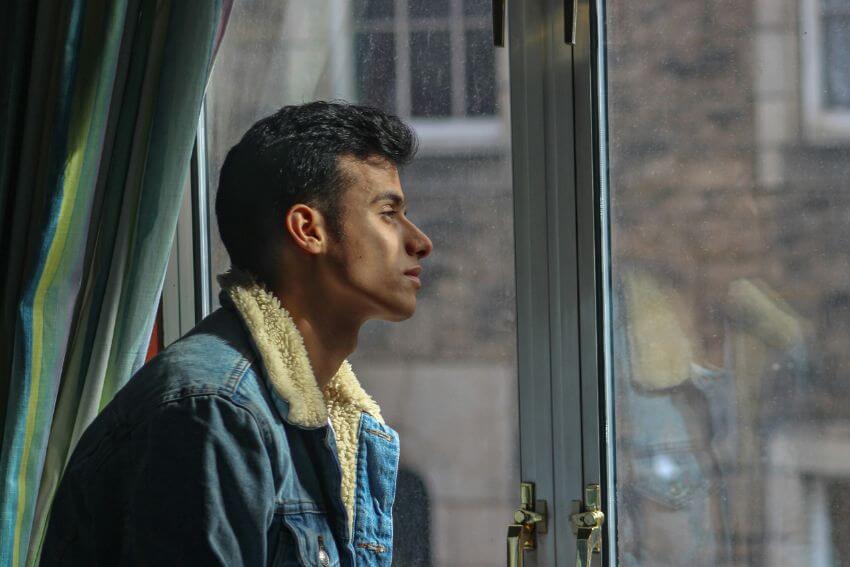 A young man gazing out a window in quiet thought, pausing to reflect on a difficult moment.