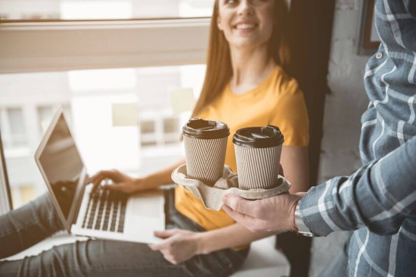 A person offering two cups of coffee to a colleague working at a laptop, the kind of small ordinary gesture an abundance mindset makes second nature.