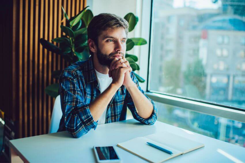 A man sitting at a desk with his hands clasped, gazing out the window in quiet thought, pausing to ask what else is true in the moment.