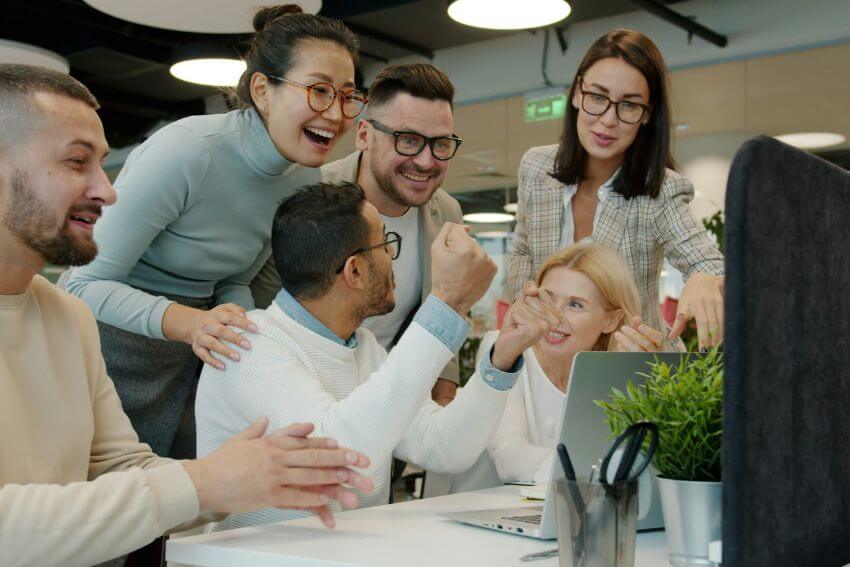 A diverse office team gathered around a laptop, cheering together over a shared win at work.