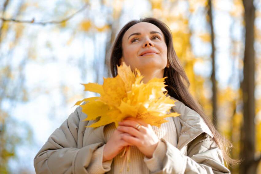 A woman holding autumn leaves and looking up with a quiet smile, naturally finding what attentional positivity asks you to notice in an ordinary moment.