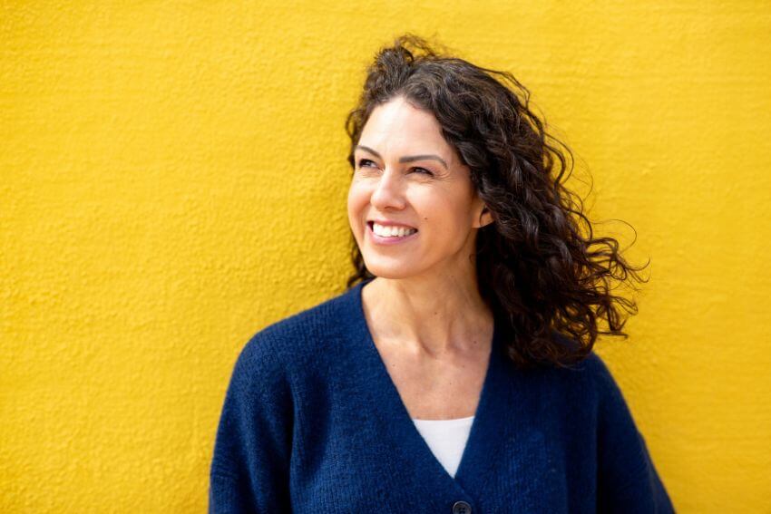 A woman smiling warmly and looking away against a bright yellow wall, radiating the quiet confidence that comes from knowing how to build trust in a relationship.