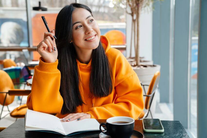 A woman smiling and writing in her notebook at a cafe, pausing to think about the people who helped her get there.