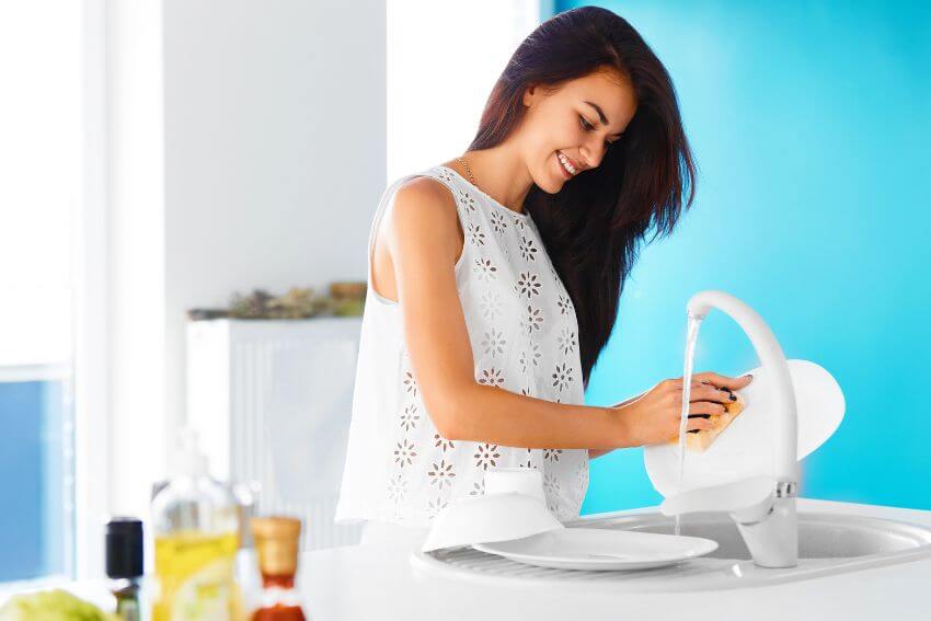A woman smiling while washing dishes at a bright kitchen sink, the everyday moment that attentional positivity is made of.