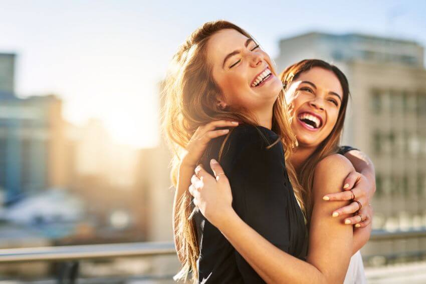 Two women laughing and hugging each other in warm sunlight, the kind of genuine connection that comes from knowing how to build trust in a relationship over time.