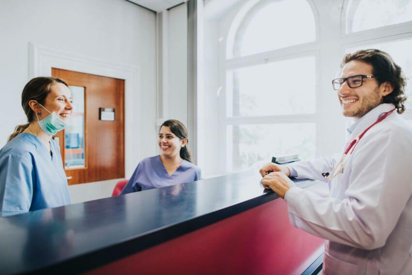 A doctor and two nurses sharing a genuine laugh at the nurses station, the kind of warm connection that people who are caring too much naturally create around them.