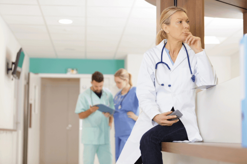 A female doctor sitting apart in a hospital corridor deep in thought, the look of someone who understands the cost of caring too much in a system built for indifference.