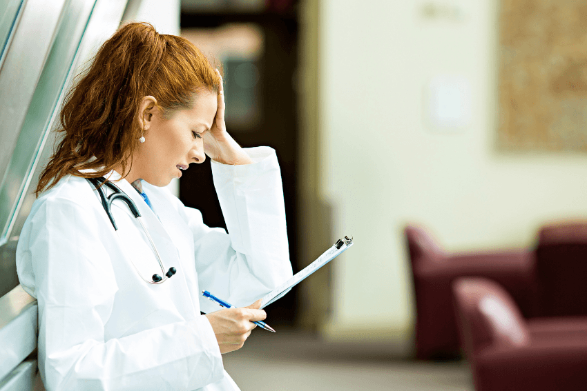 A female doctor leaning against a wall with her hand to her head, the quiet weight of caring too much inside a system that doesn't know what to do with it.